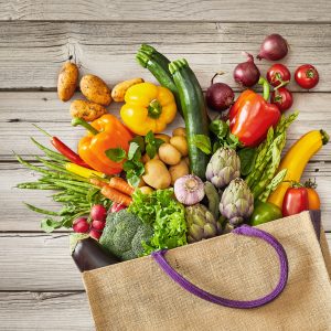 Grocery bag with purple handle that is spilling vegetables while on top of rustic wooden bench background